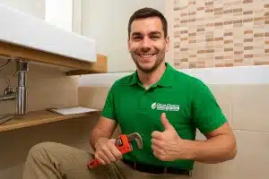 A smiling Victoria plumber from Clear Choice Plumbing kneeling under a bathroom sink, holding a pipe wrench and giving a thumbs-up — representing trusted residential plumber services in Victoria BC.